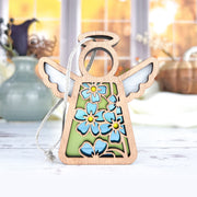 The Forget-Me-Not Ornament by Forged Flare® is a 3.5" wooden Mother's Angels® figurine with white wings and blue flowers, perfect as a charming Christmas tree ornament or flower-themed gift. Blurred vases and sunlight complete the scene.