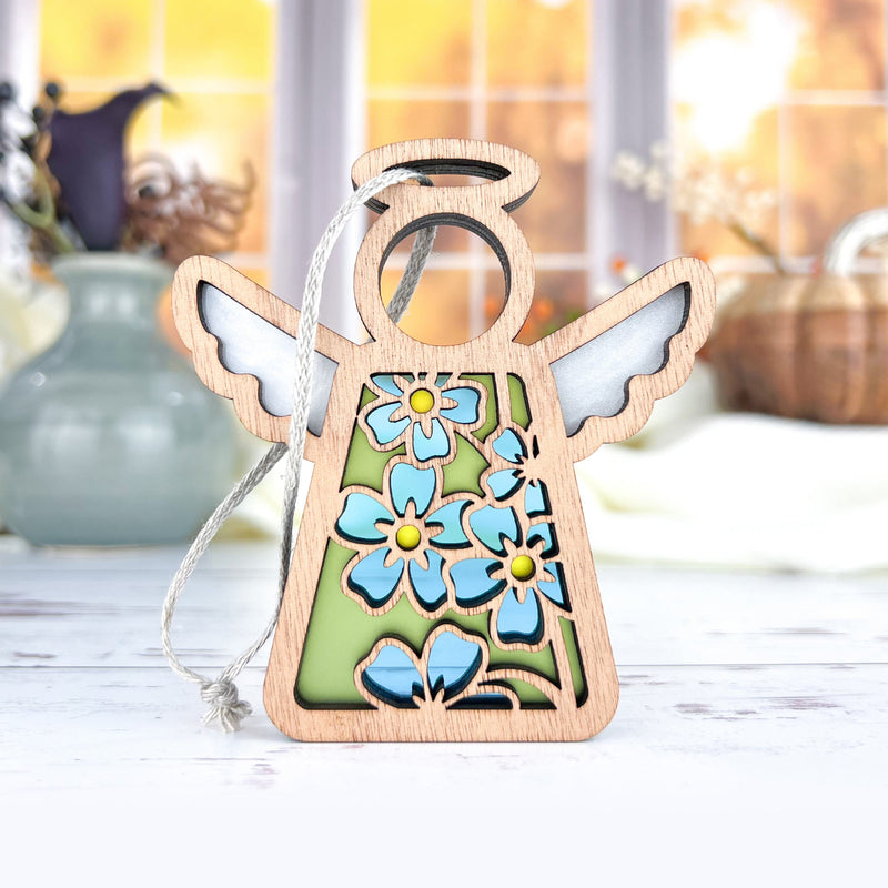 The Forget-Me-Not Ornament by Forged Flare® is a 3.5" wooden Mother's Angels® figurine with white wings and blue flowers, perfect as a charming Christmas tree ornament or flower-themed gift. Blurred vases and sunlight complete the scene.