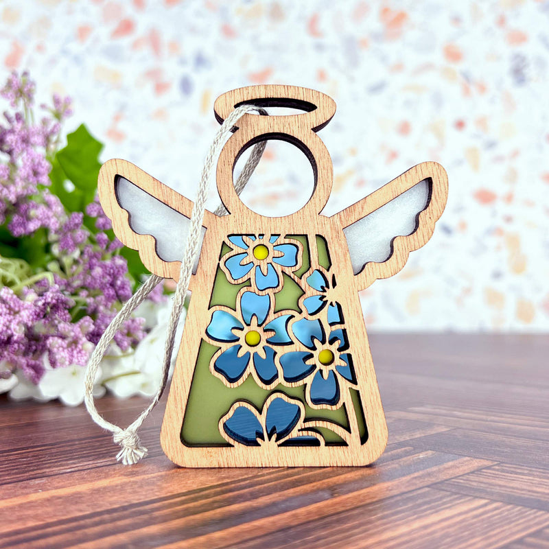 The Forget-Me-Not Ornament by Forged Flare® is a 3.5" wooden angel figurine with floral cutouts and blue flowers, perfect for gifting or as a Christmas tree ornament. Comes with a silver hanging string. From the Mother's Angels® collection.