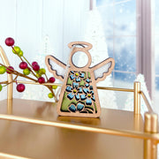 The Forget-Me-Not Ornament, a 3.5" angel figurine from Mother's Angels® by Forged Flare®, stands on a gold shelf with red and green berries, snowy trees, and a frosty window in the background.