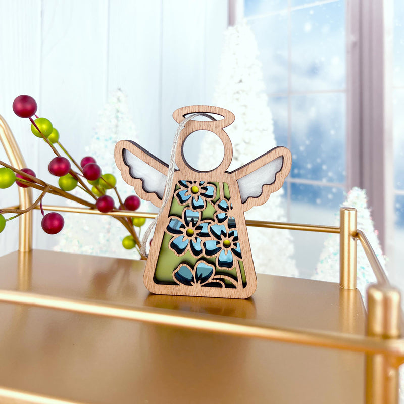 The Forget-Me-Not Ornament, a 3.5" angel figurine from Mother's Angels® by Forged Flare®, stands on a gold shelf with red and green berries, snowy trees, and a frosty window in the background.