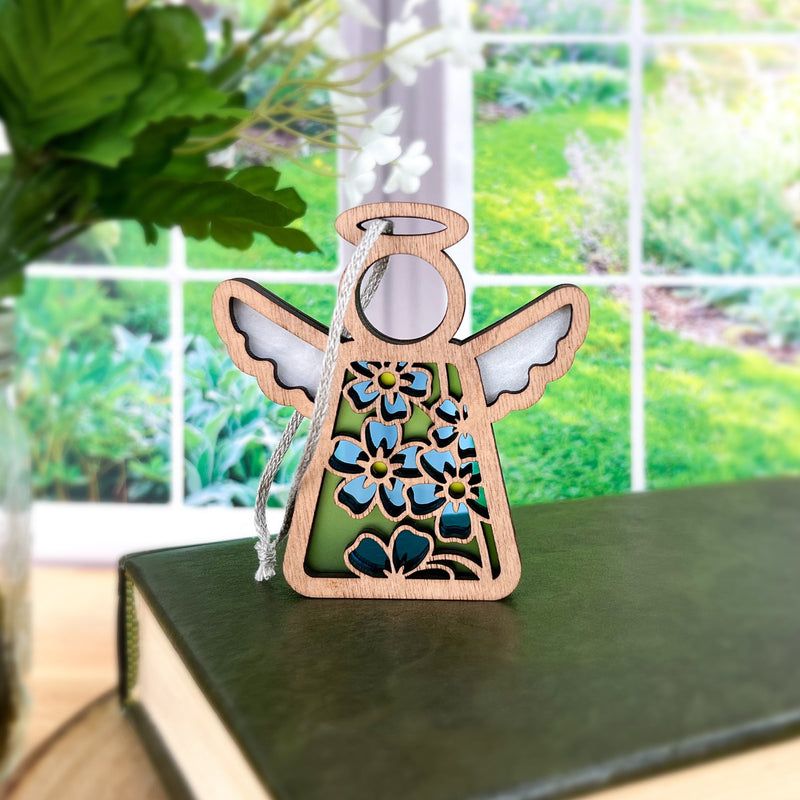 The Forget-Me-Not Ornament by Forged Flare®, a 3.5" angel figurine from the Mother's Angels® collection, features white wings and floral cut-out patterns. It stands on a closed book against a bright window, with its round halo glowing softly. The green foliage visible through the window creates a natural, peaceful background that beautifully complements the delicate forget-me-not flowers.