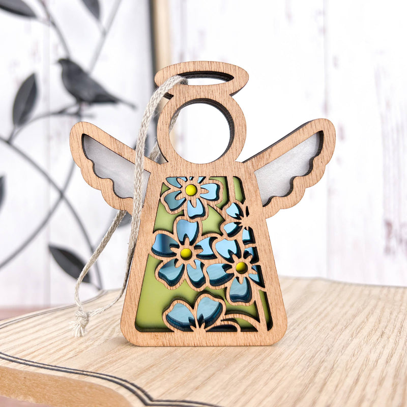 The Forged Flare® Forget-Me-Not Ornament is a 3.5" Mother's Angels® wooden angel figurine with blue flowers and green accents, hanging by a string—an ideal Christmas tree ornament and thoughtful flower gift. A metal bird wall decor appears blurred in the background.