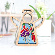 The Forged Flare® Fuchsia Ornament is a 3.5" Mother's Angels® figurine with stained glass-style wings and a fuchsia flower, standing on a white surface—perfect as a Christmas tree ornament or flower gift. Blurred plants and desk items are in the background.