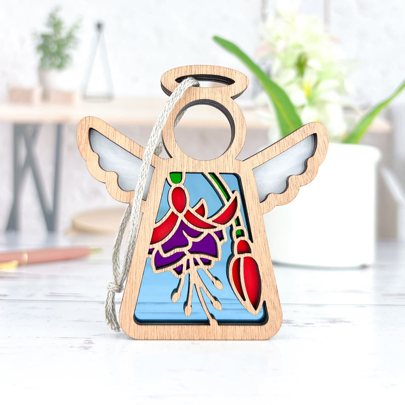 The Forged Flare® Fuchsia Ornament is a 3.5" Mother's Angels® figurine with stained glass-style wings and a fuchsia flower, standing on a white surface—perfect as a Christmas tree ornament or flower gift. Blurred plants and desk items are in the background.