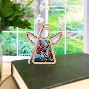 A Forged Flare® Mother's Angels® Fuchsia Ornament, a 3.5" angel figurine, rests on a closed green book by the window with garden views and leafy greens—suggesting an ideal flower-inspired gift.