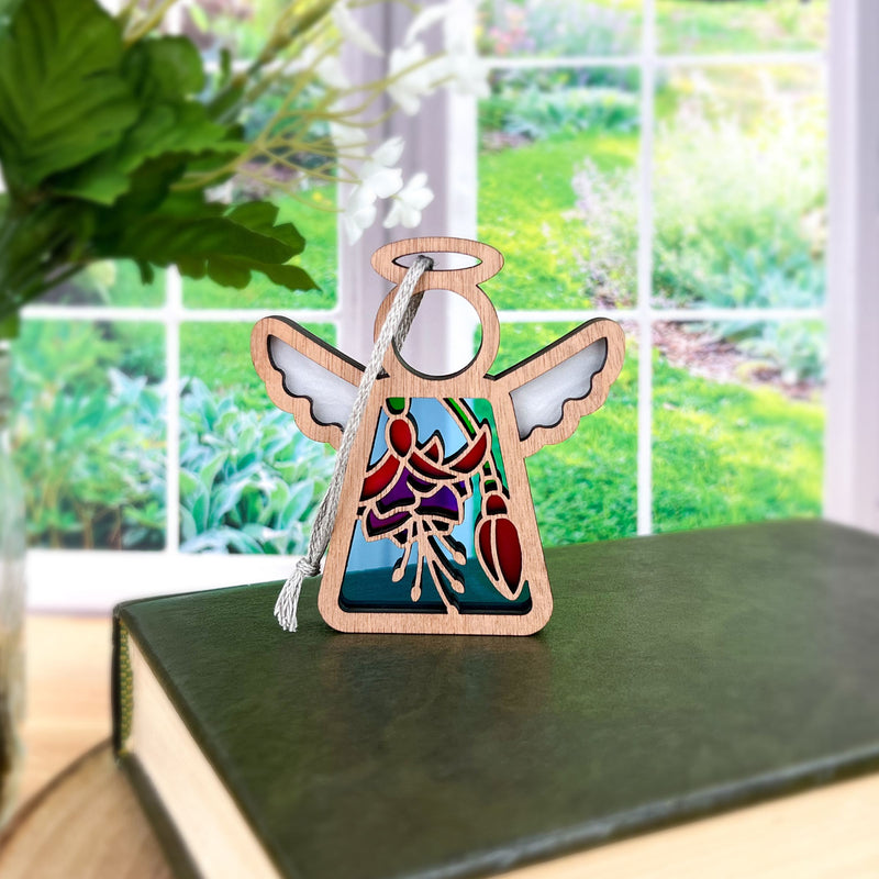 A Forged Flare® Mother's Angels® Fuchsia Ornament, a 3.5" angel figurine, rests on a closed green book by the window with garden views and leafy greens—suggesting an ideal flower-inspired gift.