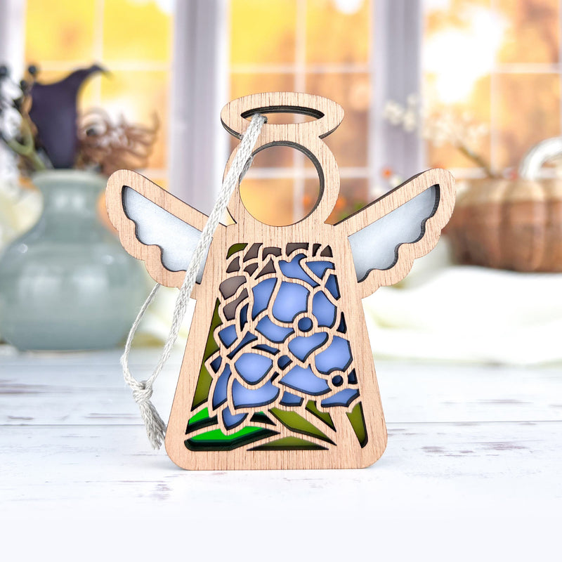The Forged Flare® Hydrangea Ornament is a 3.5" Mother's Angels® figurine with white wings and a blue hydrangea design, standing on a light surface with vases and bright windows softly blurred in the background.