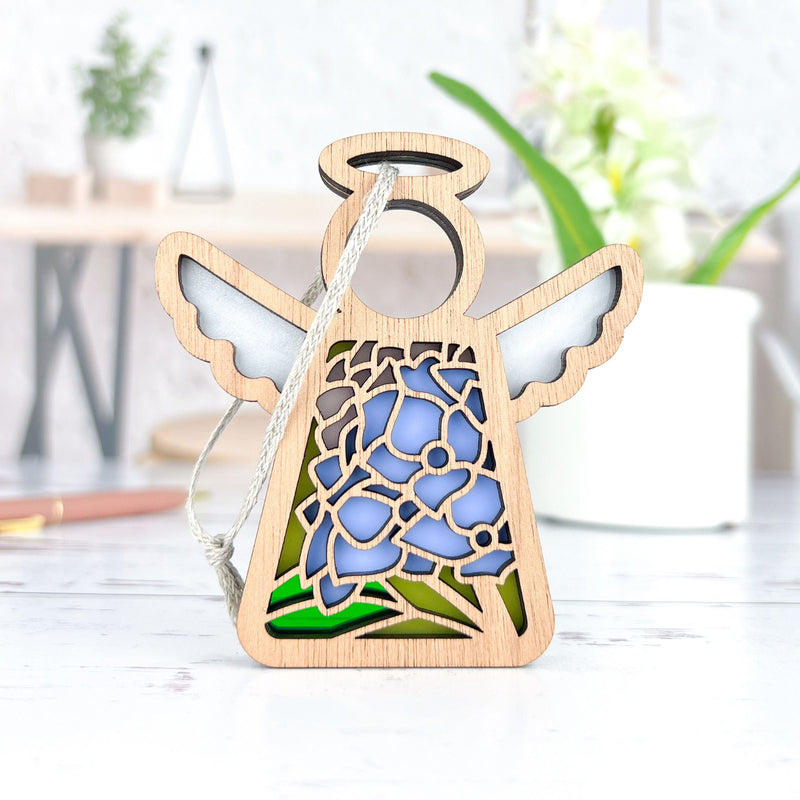 The Forged Flare® Mother's Angels® Hydrangea Ornament is a 3.5" wooden angel figurine with white wings, a halo, and blue hydrangea flower cutouts, plus a hanging string—shown against a blurred indoor background with plants.