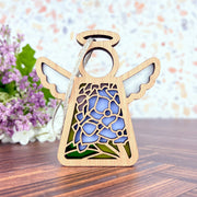 A Forged Flare® Mother's Angels® Hydrangea Ornament, a 3.5" wooden angel figurine with blue and green stained glass-style hydrangea design, stands on wood with blurred purple flowers and greenery in the background.