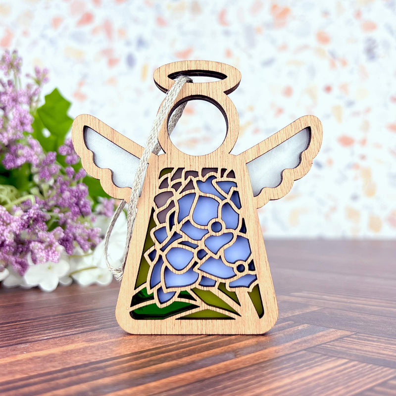 A Forged Flare® Mother's Angels® Hydrangea Ornament, a 3.5" wooden angel figurine with blue and green stained glass-style hydrangea design, stands on wood with blurred purple flowers and greenery in the background.