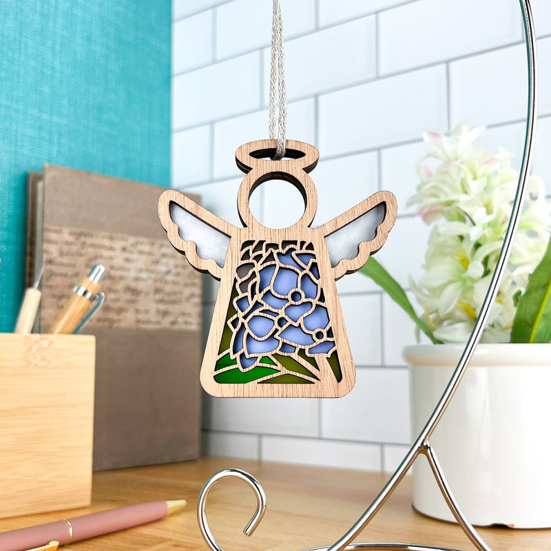 The Forged Flare® Hydrangea Ornament is a 3.5" Mother's Angels® wooden angel figurine decorated with blue hydrangea and green leaves, perfect as a Christmas ornament or decor near your desk.