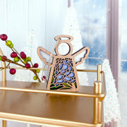 The Forged Flare® Hydrangea Ornament, a 3.5" Mother's Angels® angel figurine, is displayed on a gold shelf among frosted white trees and red berries, with snow-kissed windows in the background.