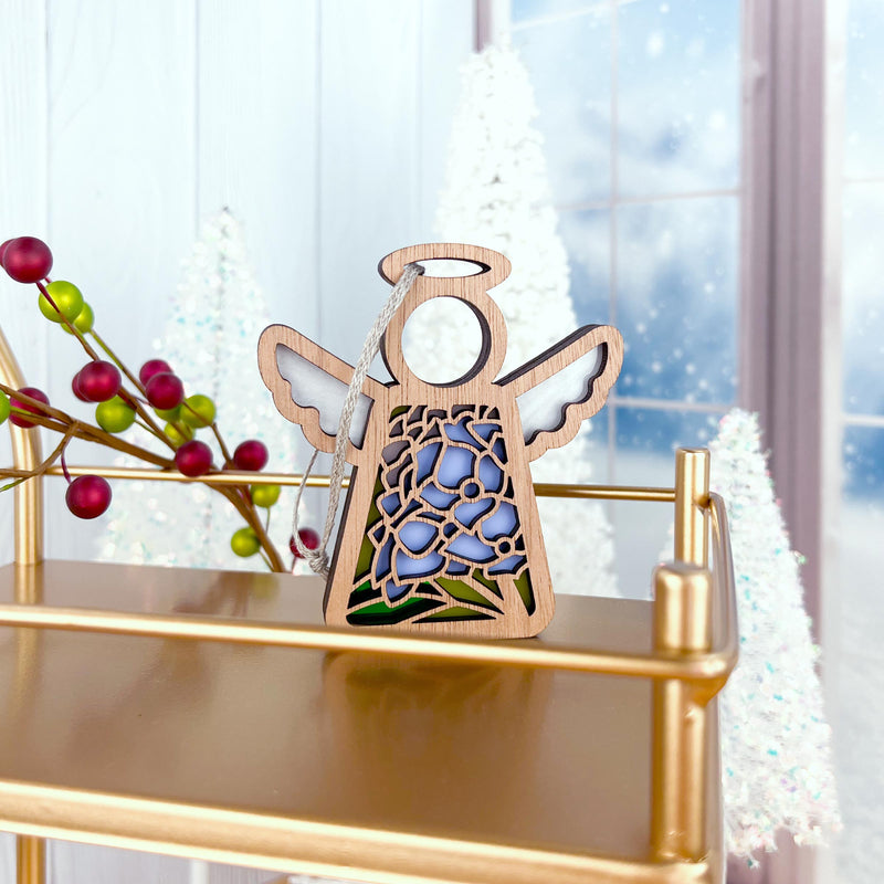 The Forged Flare® Hydrangea Ornament, a 3.5" Mother's Angels® angel figurine, is displayed on a gold shelf among frosted white trees and red berries, with snow-kissed windows in the background.