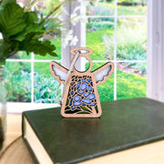 The Forged Flare® Hydrangea Ornament, a 3.5" Mother's Angels® angel figurine with blue hydrangea details, sits on a closed green book against a bright window and garden backdrop.