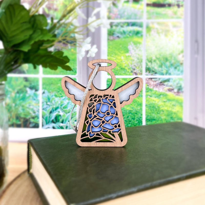 The Forged Flare® Hydrangea Ornament, a 3.5" Mother's Angels® angel figurine with blue hydrangea details, sits on a closed green book against a bright window and garden backdrop.