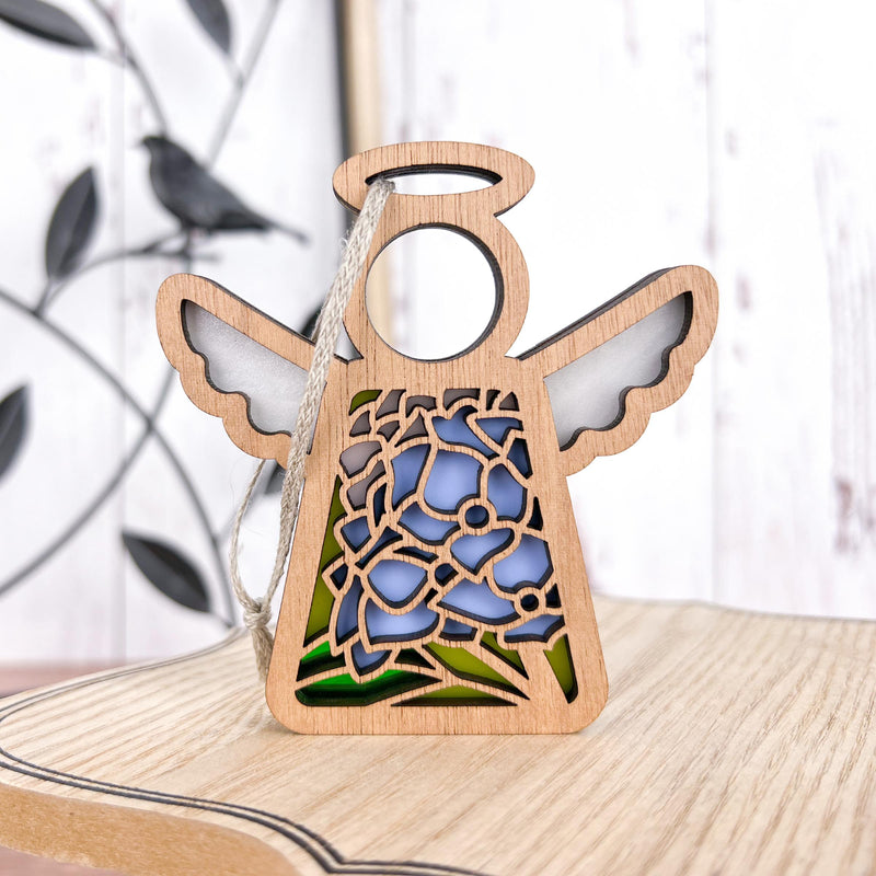 A 3.5" wooden angel figurine with intricate floral cutouts, part of the Mother's Angels® collection from Forged Flare®, reminiscent of the Hydrangea Ornament, stands on a light wooden surface. The floral design in the angel's dress is highlighted with blue and green stained glass. In the blurred background, a metal branch with a bird silhouette is visible.