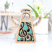 The Forged Flare® Music Ornament, a 3.5" Mother's Angels® angel figurine with blue accents and black musical notes, stands on a white surface—an ideal gift for music teachers. Blurred plants and desk items appear in the background.