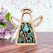 The Forged Flare® Mother's Angels® Music Ornament is a 3.5" wooden angel with a musical note design, white wings, and a silver hanging string—an ideal music teacher gift. Shown on wood with purple flowers in the background.