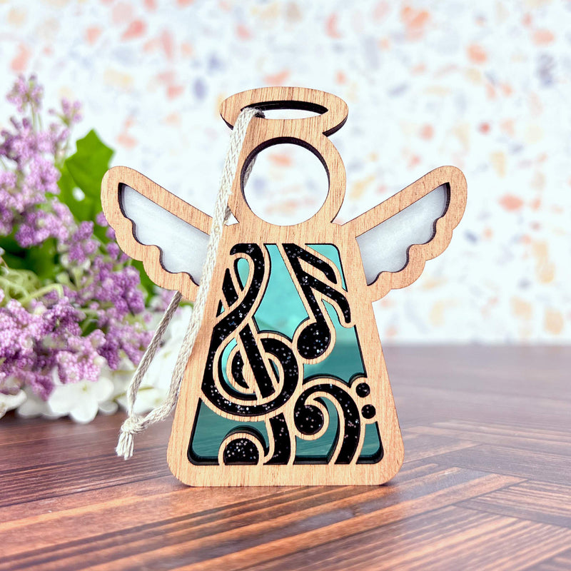 The Forged Flare® Mother's Angels® Music Ornament is a 3.5" wooden angel with a musical note design, white wings, and a silver hanging string—an ideal music teacher gift. Shown on wood with purple flowers in the background.