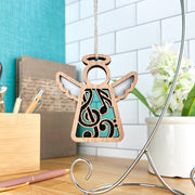 The Forged Flare® Mother's Angels® 3.5" wooden angel music ornament with musical notes and a treble clef cutout hangs on a silver stand, creating a thoughtful desk gift for music teachers.