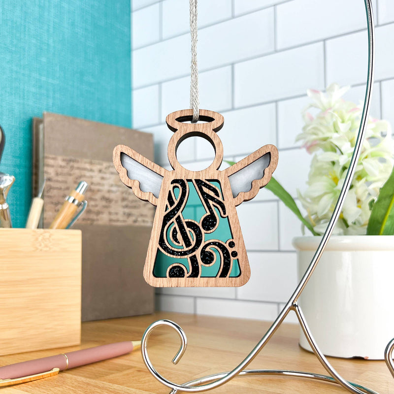 The Forged Flare® Mother's Angels® 3.5" wooden angel music ornament with musical notes and a treble clef cutout hangs on a silver stand, creating a thoughtful desk gift for music teachers.