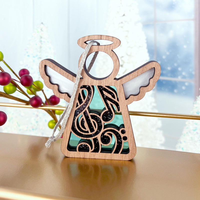 The Forged Flare® Music Ornament is a 3.5" (H) Mother's Angels® wooden angel figurine with a cut-out musical note, standing on gold near festive decor—white trees, red berries, and a blurred wintery window. Perfect as a music teacher gift.