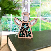 The Forged Flare® Mother's Angels® Music Ornament (3.5" tall), featuring intricate black and white designs and a silver string, rests on a green book—an ideal gift for music teachers. A bright garden is visible outside the window.