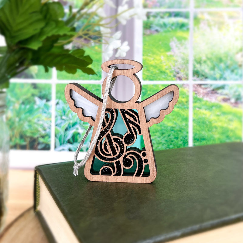 The Forged Flare® Mother's Angels® Music Ornament (3.5" tall), featuring intricate black and white designs and a silver string, rests on a green book—an ideal gift for music teachers. A bright garden is visible outside the window.