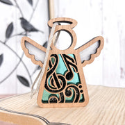 The Forged Flare® Mother's Angels® 3.5" Music Ornament features an angel figurine with white wings, a halo, and black and teal musical notes & swirls—ideal for gifting or as a music teacher keepsake. A blurred metal bird sits in the background.