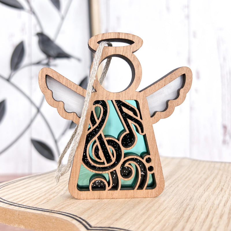 The Forged Flare® Mother's Angels® 3.5" Music Ornament features an angel figurine with white wings, a halo, and black and teal musical notes & swirls—ideal for gifting or as a music teacher keepsake. A blurred metal bird sits in the background.