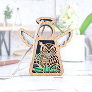 The Forged Flare® Mother's Angels® Owl Ornament is a 3.5" wooden angel figurine with wings, perfect for bird lovers. It hangs by a string and features a detailed carved owl, shown on a white surface with blurred plants in the background.