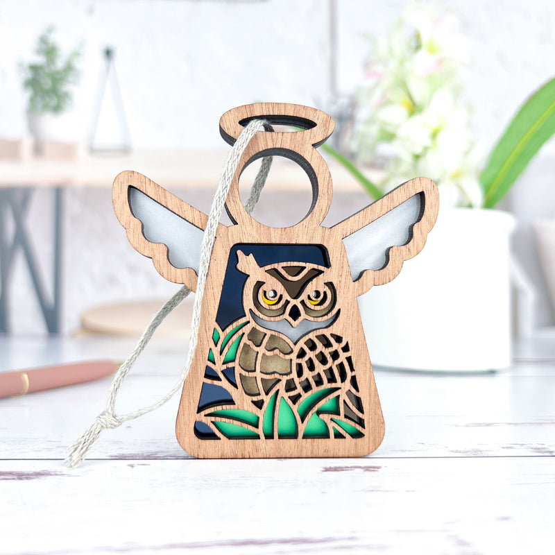 The Forged Flare® Mother's Angels® Owl Ornament is a 3.5" wooden angel figurine with wings, perfect for bird lovers. It hangs by a string and features a detailed carved owl, shown on a white surface with blurred plants in the background.