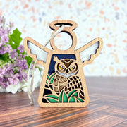 The Forged Flare® Mother's Angels® Owl Ornament is a 3.5" wooden angel figurine with outstretched wings, green accents, and a carved owl design. Ideal for bird lovers, it hangs by a string. Purple flowers appear blurred in the background.