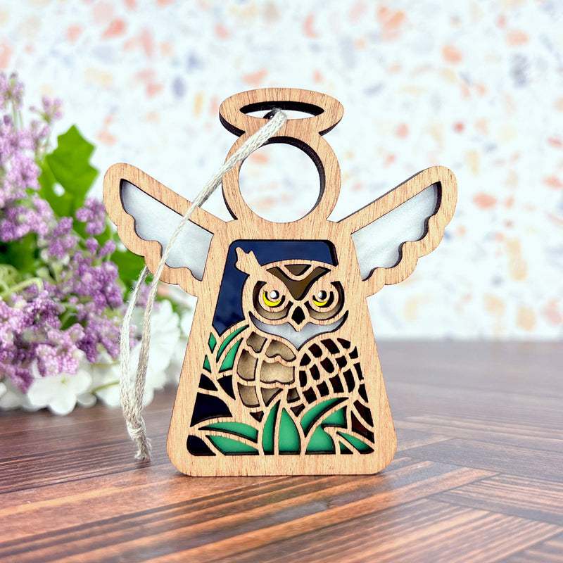 The Forged Flare® Mother's Angels® Owl Ornament is a 3.5" wooden angel figurine with outstretched wings, green accents, and a carved owl design. Ideal for bird lovers, it hangs by a string. Purple flowers appear blurred in the background.