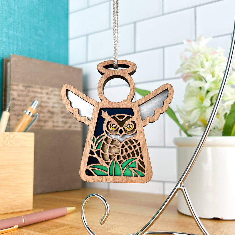 The Forged Flare® Owl Ornament | 3.5" (H) Angel Figurine from the Mother's Angels® line hangs indoors on a metal stand, with a desk and plant in the background—a charming bird lover gift for any nature enthusiast.