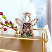 The Forged Flare® Owl Ornament (Mother’s Angels®, 3.5" Angel Figurine) sits on a gold shelf with frosted trees, a window, and festive berries—perfect winter decor and a thoughtful bird lover gift.