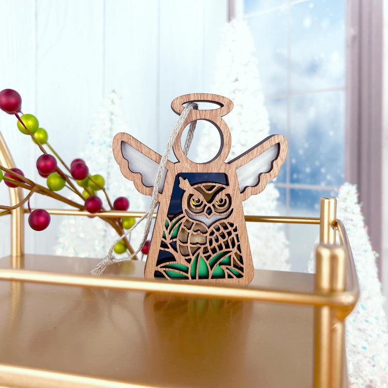The Forged Flare® Owl Ornament (Mother’s Angels®, 3.5" Angel Figurine) sits on a gold shelf with frosted trees, a window, and festive berries—perfect winter decor and a thoughtful bird lover gift.