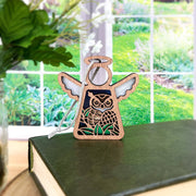 The Forged Flare® Owl Ornament is a 3.5" Mother’s Angels® angel figurine with wings and white ribbon, perfect as a charming bird lover or bird watching gift. It stands beautifully near a window on a closed green book.