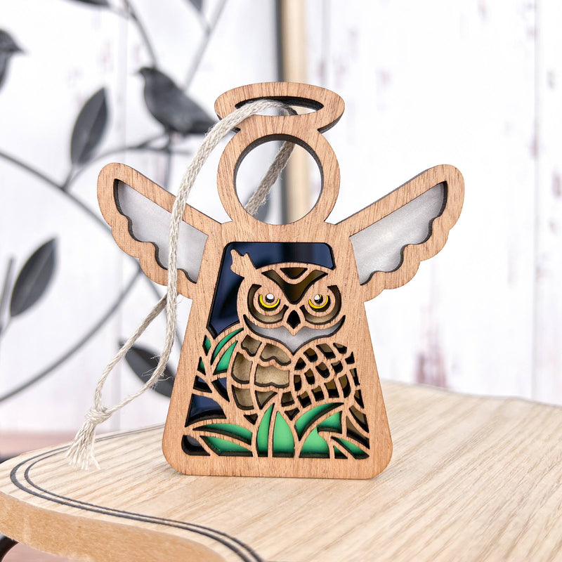 The Forged Flare® Owl Ornament is a 3.5" Mother's Angels® angel figurine with a cut-out owl and green grass. It comes with a hanging string and rests on wood, making it a perfect gift for bird lovers or bird watchers.