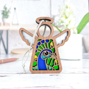 The Peacock Ornament by Forged Flare® is a 3.5" Mother's Angels® angel figurine with white wings and a stained-glass style blue and green peacock—an ideal bird lover gift, shown on a white tabletop with blurred plants in the background.