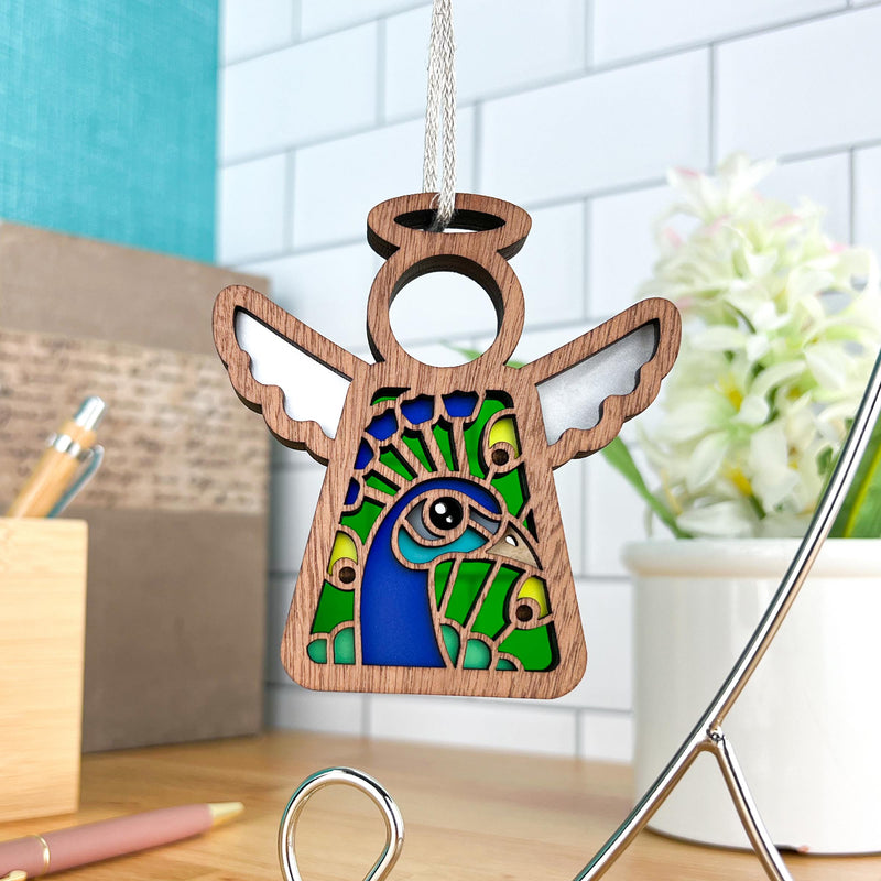 The Forged Flare® Peacock Ornament, a 3.5" Mother's Angels® wooden angel figurine with a peacock design, hangs by a silver string—ideal for bird lovers and shown against white subway tiles, flowers, and pens on a desk.
