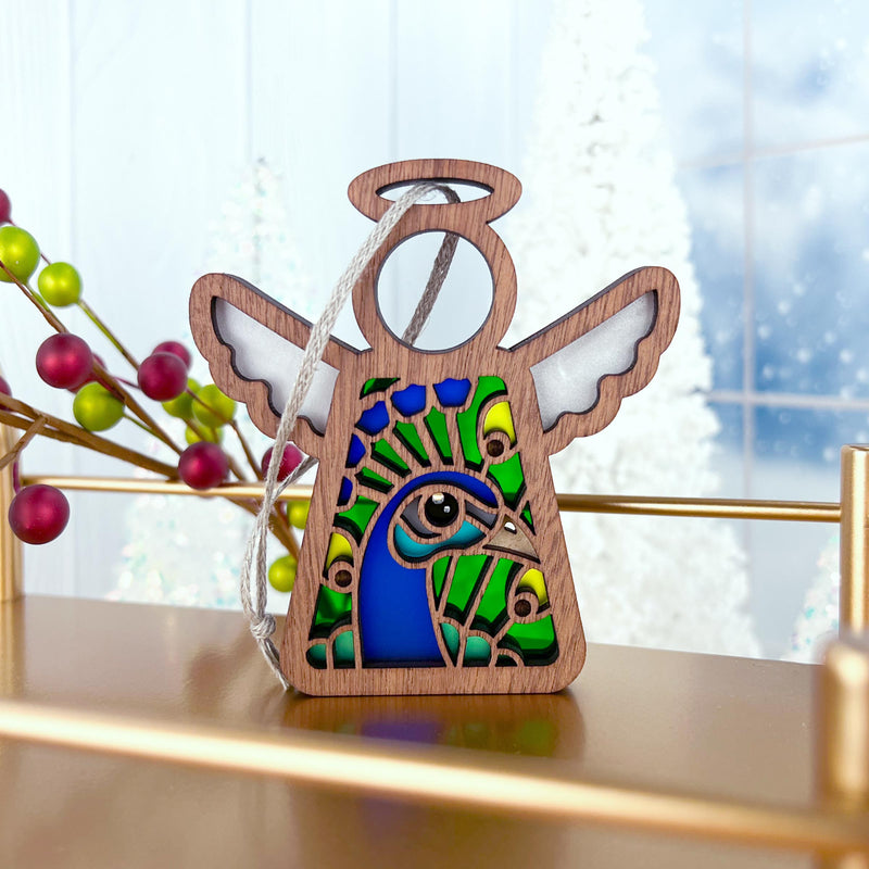 The Forged Flare® Peacock Ornament is a 3.5" wooden Mother's Angels® figurine with white wings and a blue, green, and yellow stained-glass-style peacock design. It hangs by a white string, perfect for holiday décor.