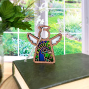 The Forged Flare® Peacock Ornament, a 3.5" stained-glass angel figurine from Mother's Angels®, stands on a closed green book by a bright window with a white hanging string, and a plant is partly visible on the left—ideal for bird lovers.
