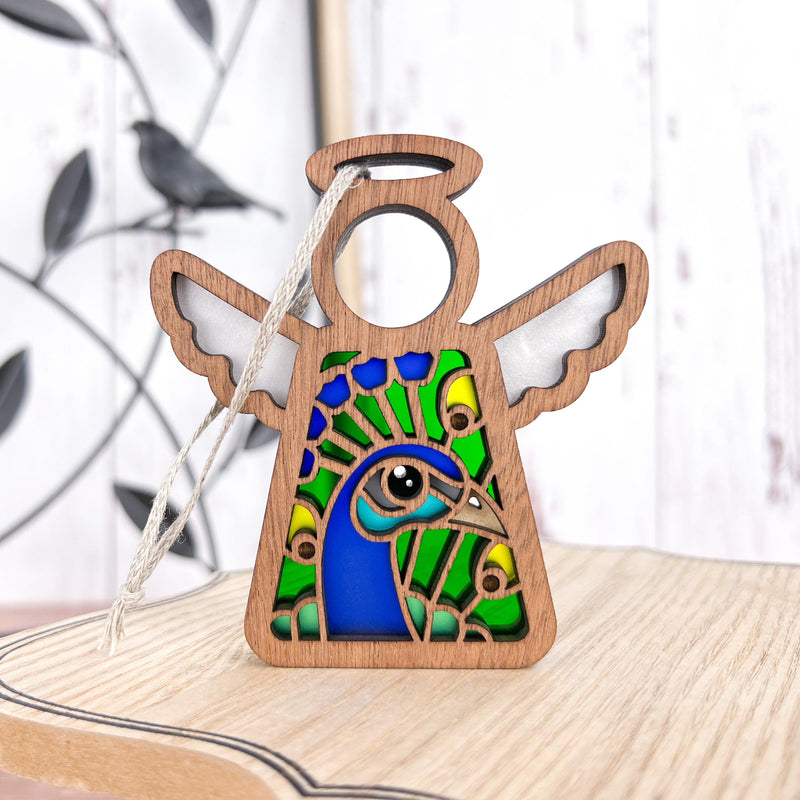 The Forged Flare® Peacock Ornament | 3.5" (H) Angel Figurine from Mother's Angels® shines in vibrant blues, greens, and yellows on a light wood surface. It includes a hanging string; ideal as a bird lover gift. A blurred metal bird ornament appears in the background.
