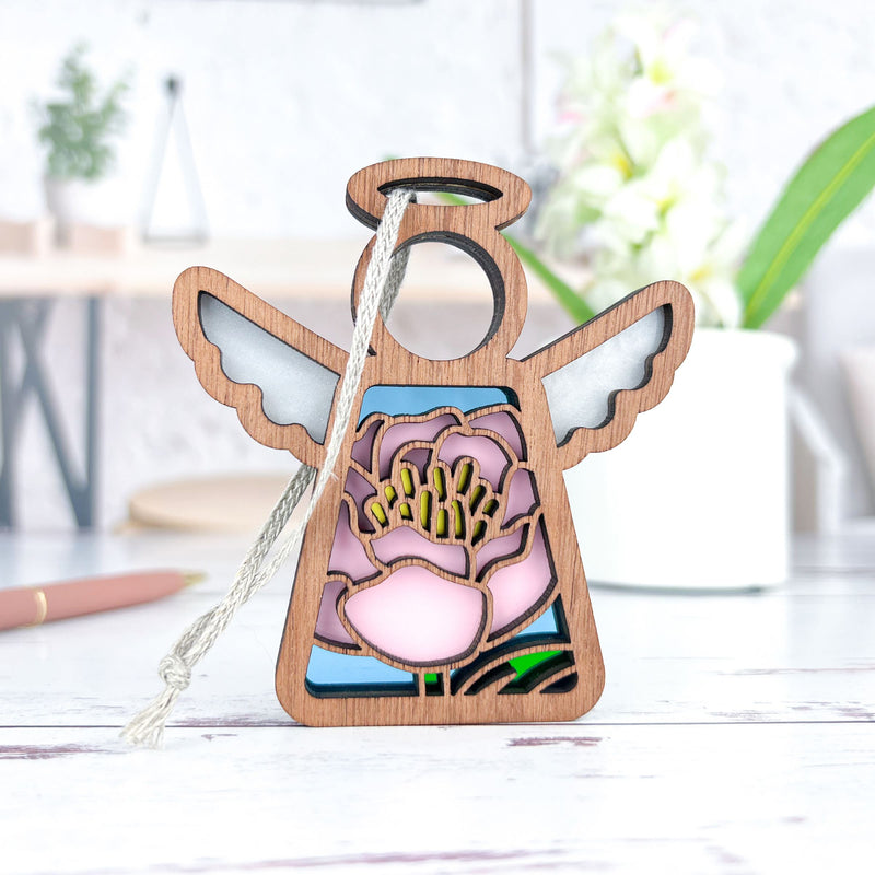 The Pink Peony Ornament by Forged Flare® is a 3.5" wooden Mother's Angels® figurine with stained glass-style wings and a pink peony center, perfect as a Christmas tree ornament. It stands on a white surface with blurred decor behind it.
