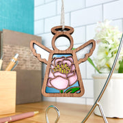 The Forged Flare® Pink Peony Ornament | 3.5" (H) Angel Figurine from Mother's Angels® features a stained glass-style peony design and hangs indoors, with a blurred background of a plant, pen, and office supplies.