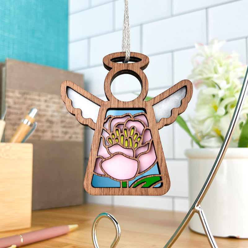 The Forged Flare® Pink Peony Ornament | 3.5" (H) Angel Figurine from Mother's Angels® features a stained glass-style peony design and hangs indoors, with a blurred background of a plant, pen, and office supplies.