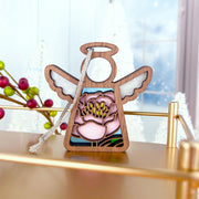 The Forged Flare® Pink Peony Ornament, a 3.5" Mother's Angels® angel figurine, sits on a gold shelf with festive red berries and white trees softly blurred in the background.