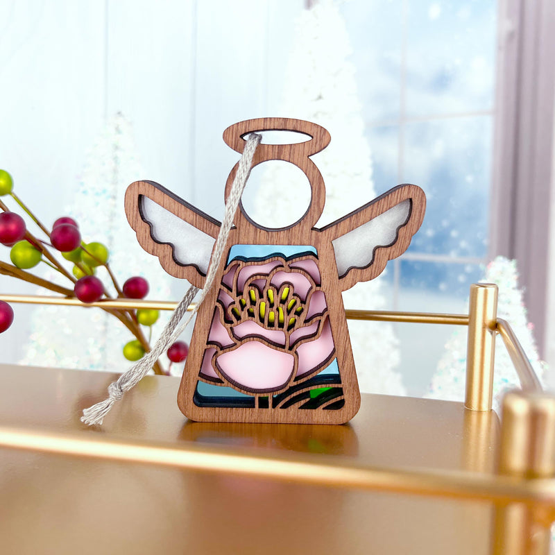The Forged Flare® Pink Peony Ornament, a 3.5" Mother's Angels® angel figurine, sits on a gold shelf with festive red berries and white trees softly blurred in the background.